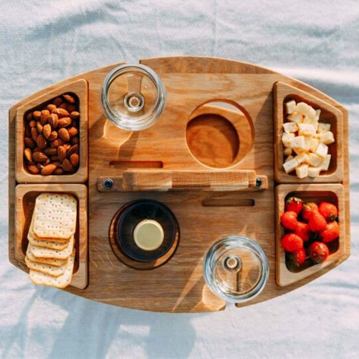 Outdoor foldable wooden wine table holding two wine glasses and snacks, perfect for a cozy picnic setup.